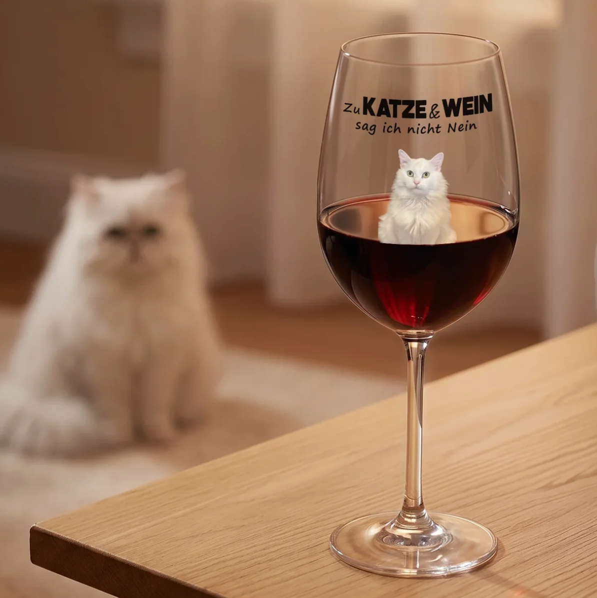 Personalisiertes Weinglas mit schwarzer Schrift und rotem Wein, daneben eine weiße Katze auf einem Holz Tisch.