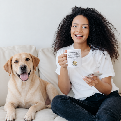 Person mit braunen, lockigen Haaren hält eine weiße Tasse mit Hundemotiv, neben einem Labrador auf dem Sofa.