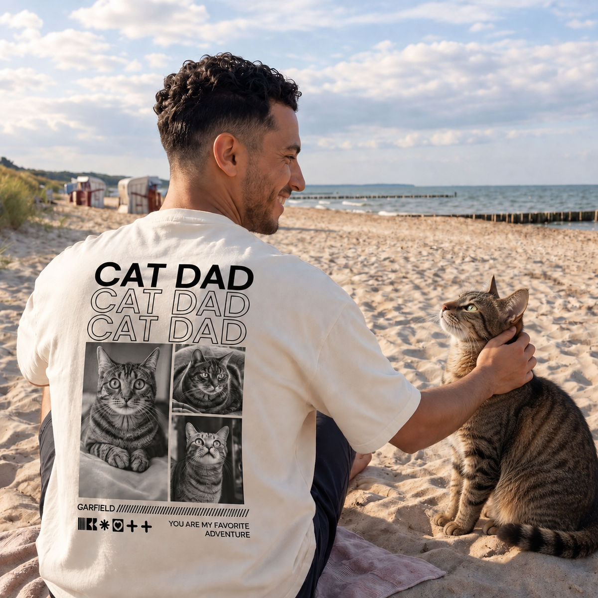 Beige T-Shirt mit Druck "Cat Dad" und Katzenmotiven auf dem Rücken, getragen von einem Mann am Strand.