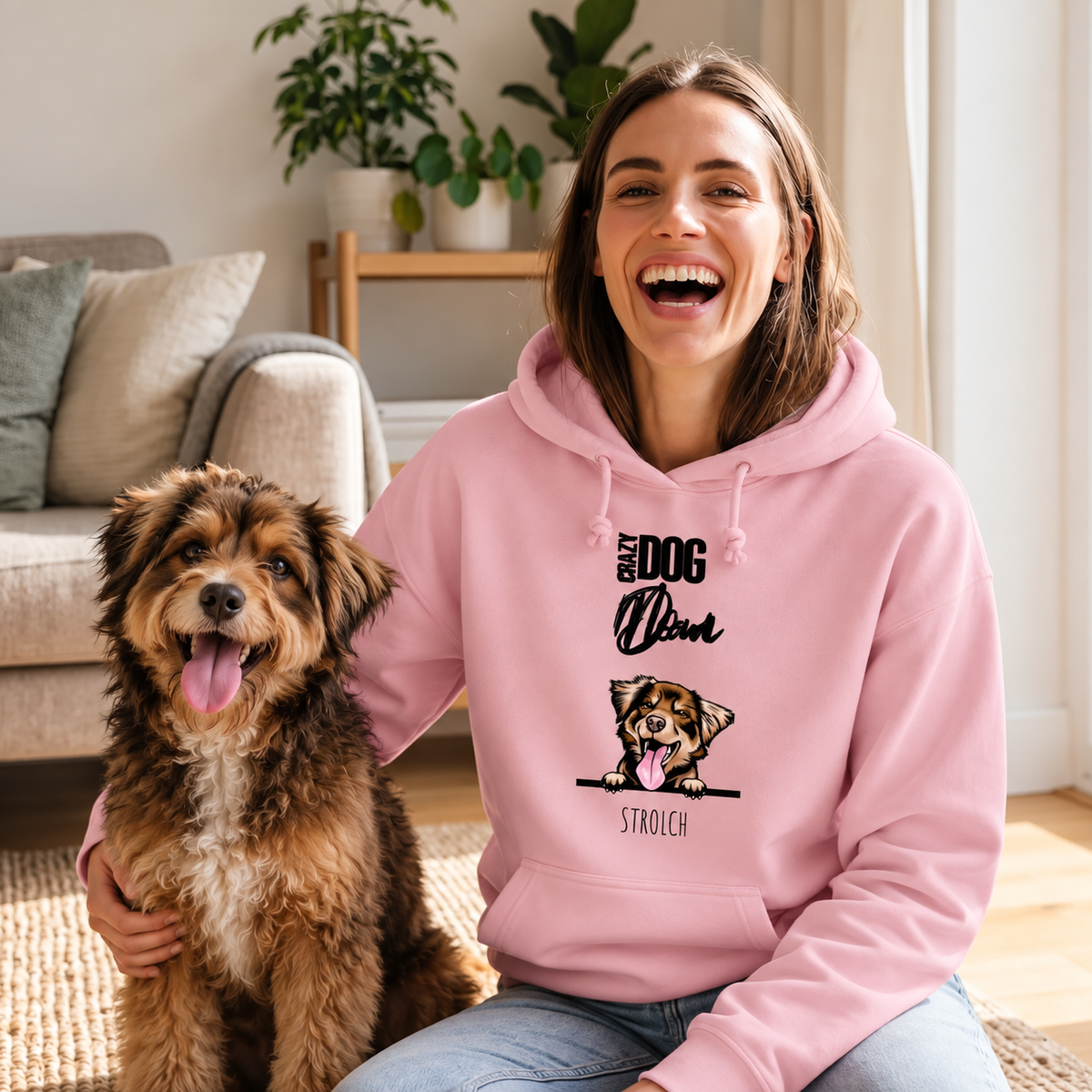 Frau mit lächelndem Gesicht trägt einen rosa Hoodie mit Hundemotiv, sitzt neben einem Hund auf einem Teppich.