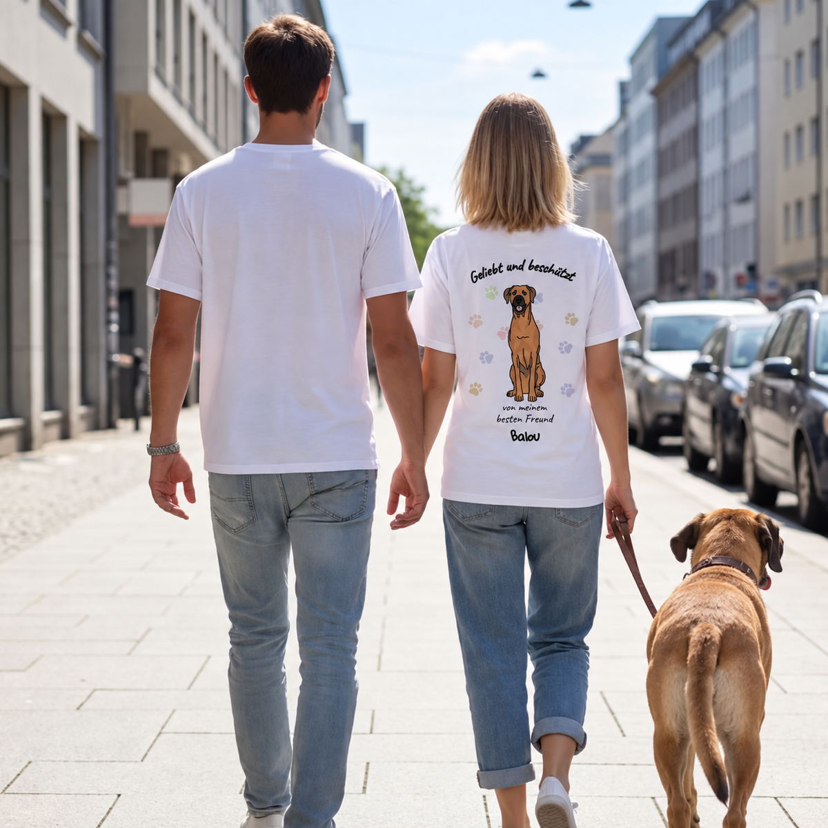 Zwei Personen tragen weiße T-Shirts mit Hundemotiv auf dem Rücken, gehen mit einem Hund spazieren.