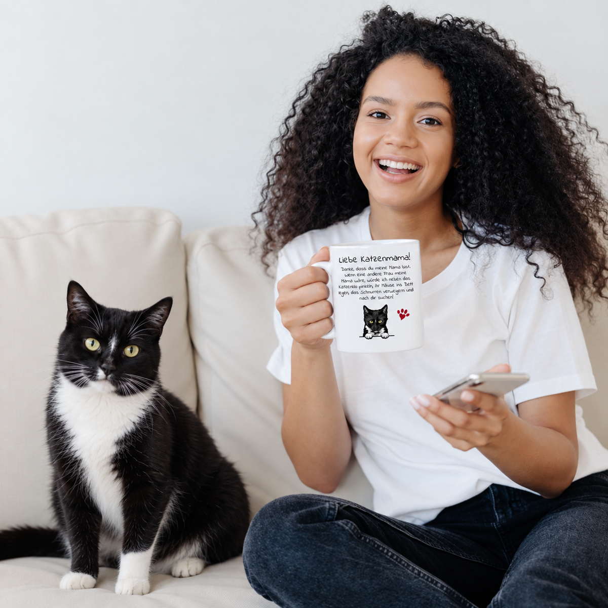 Junge Frau mit lockigem Haar hält eine personalisierte Tasse mit Katzenmotiv, schwarze Katze sitzt neben ihr auf dem Sofa.