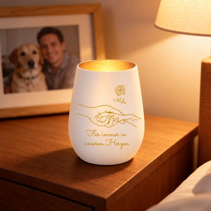 Personalisierte Trauerkerze aus Glas mit goldfarbener Schrift und Hand-Design, leuchtet sanft auf einem Nachttisch.