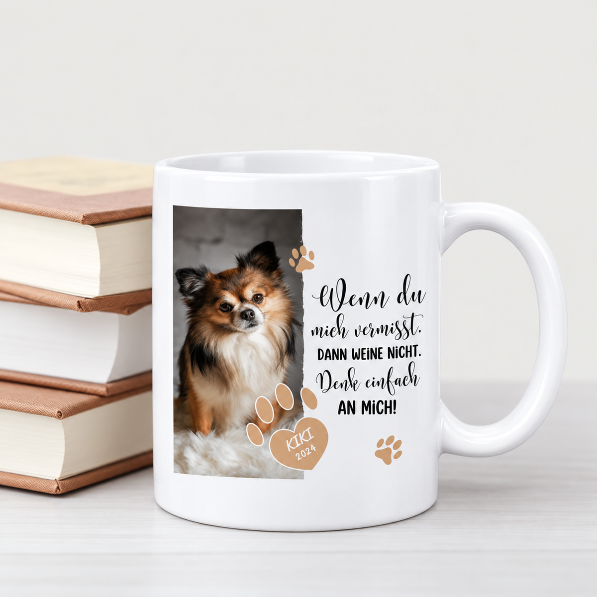 Weiße Tasse mit Aufdruck, Hundebild und Text über Vermissen, neben drei Stapeln Bücher platziert.