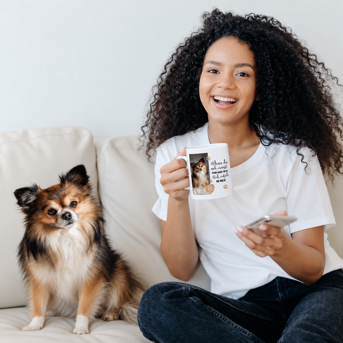 Frau mit lockigem Haar hält eine personalisierte weißer Tasse und sitzt auf einem Sofa neben einem Hund.