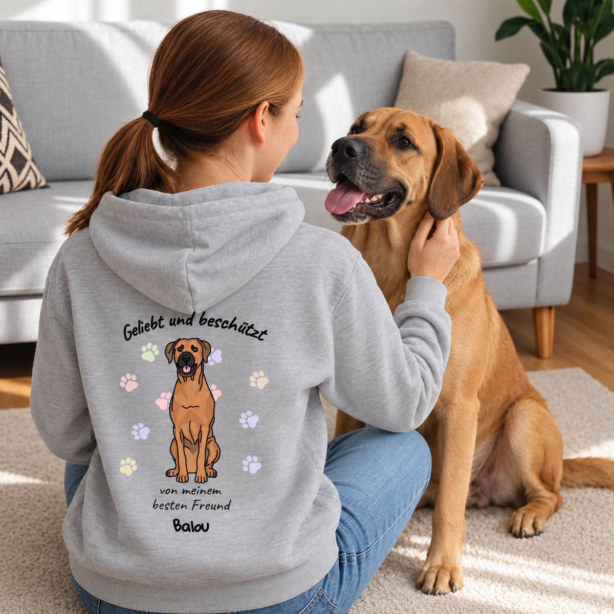 Grau Hoodie mit Hundemotiv und Text "Geliebt und beschützt" auf dem Rücken. Trägt eine Person im Sitzen.