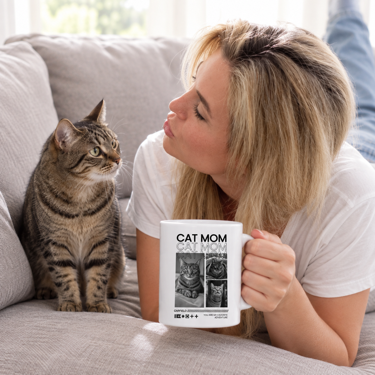 Person trinkt aus einer weißen Tasse mit der Aufschrift "CAT MOM" und sitzt neben einer Katze auf einem Sofa.