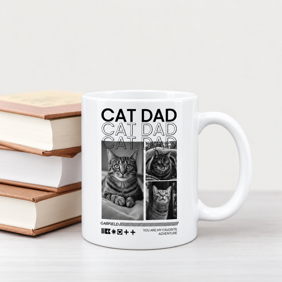 Weiße Tasse mit dem Aufdruck "CAT DAD" und Bildern von Katzen, hinter einem Stapel Bücher.