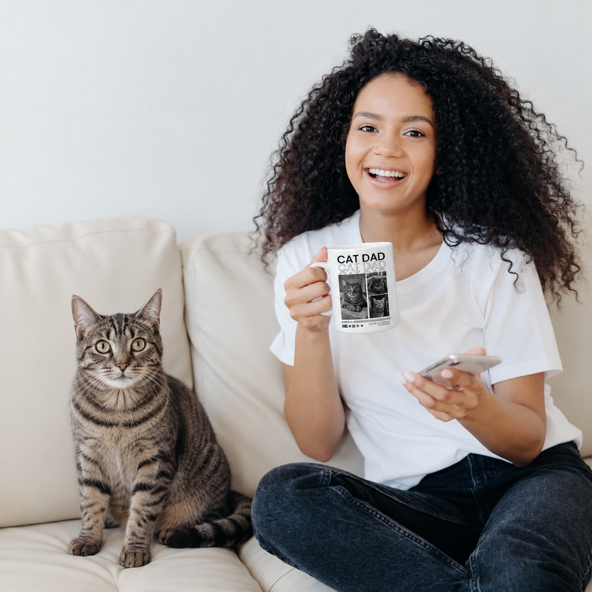Person mit lächelndem Gesicht hält eine personalisierte Tasse mit dem Aufdruck "CAT DAD" und sitzt neben einer Katze auf einem Sofa.