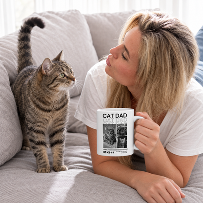 Frau sitzt auf einem Sofa und hält eine weiße Tasse mit dem Aufdruck "Cat Dad", neben ihr steht eine gestreifte Katze.
