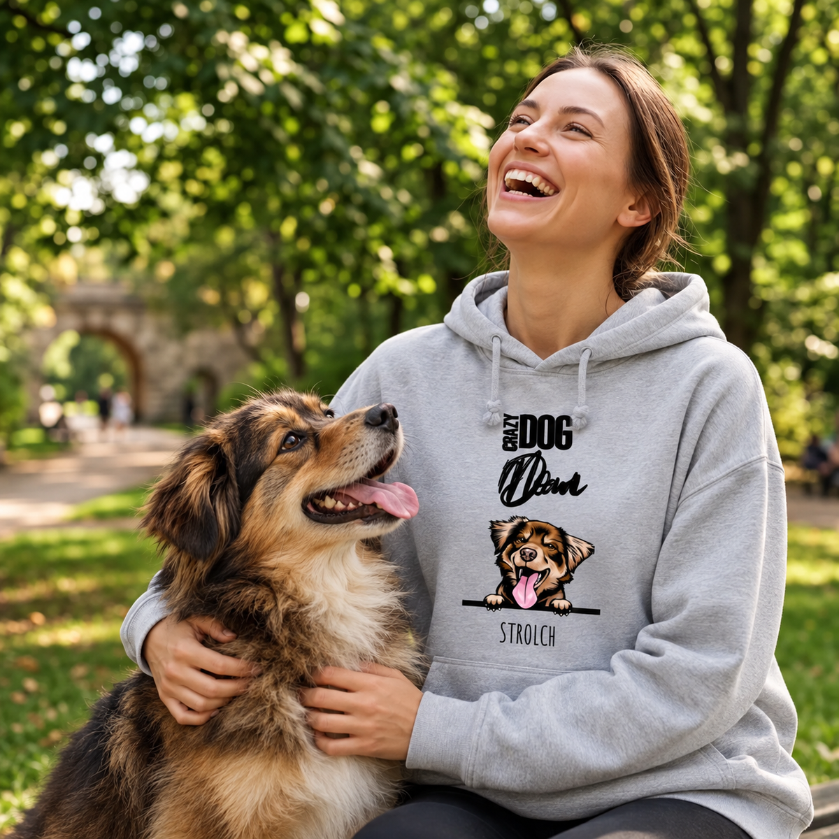 Gris-grauer Hoodie mit Hundemotiv, sitzt auf einer Frau im Park, die mit ihrem Hund spielt und lacht.
