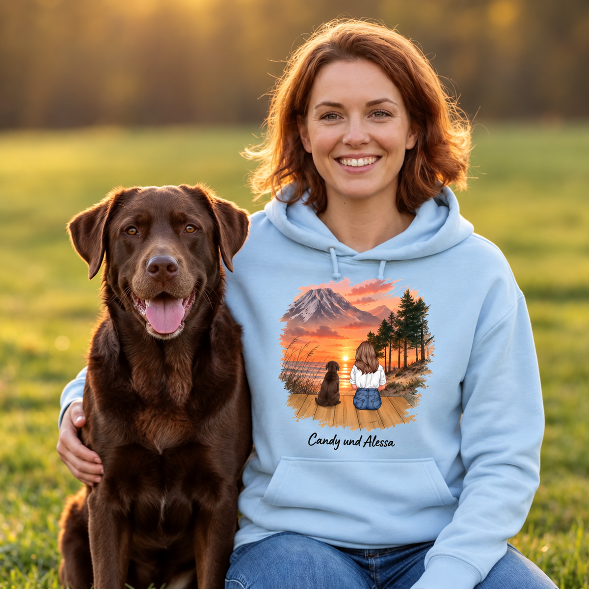 Helle, blaue Kapuzenjacke mit Grafik von Person und Hund vor einer Landschaft bei Sonnenuntergang.