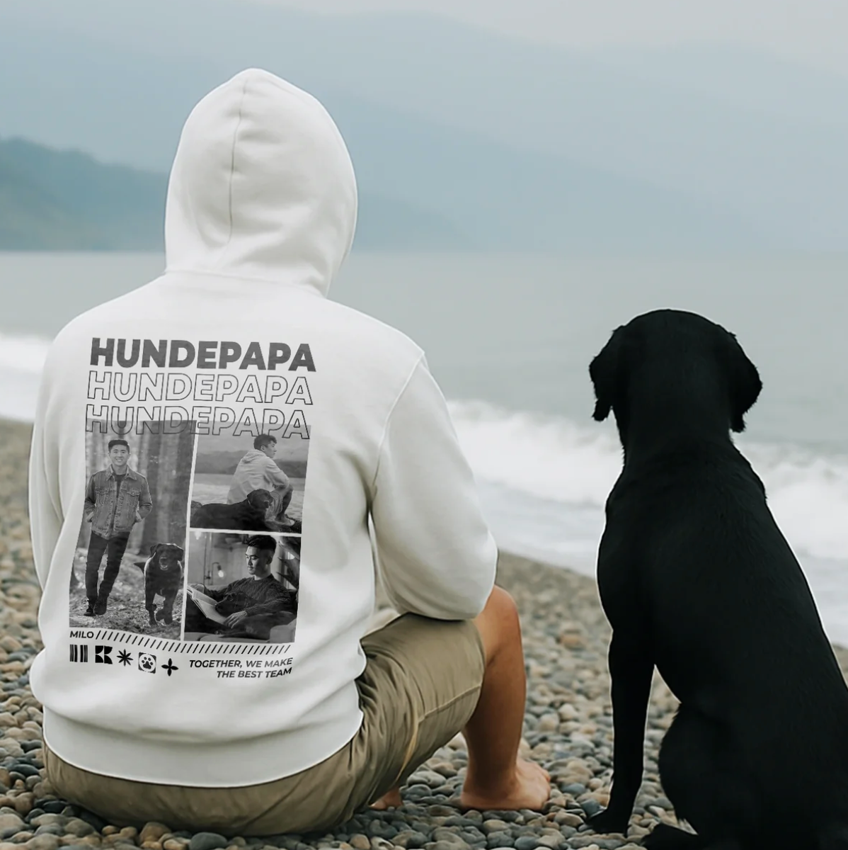 Helle Kapuzenjacke mit "Hundepapa"-Aufdruck auf der Rückseite, getragen von einer Person am Strand neben einem schwarzen Hund.