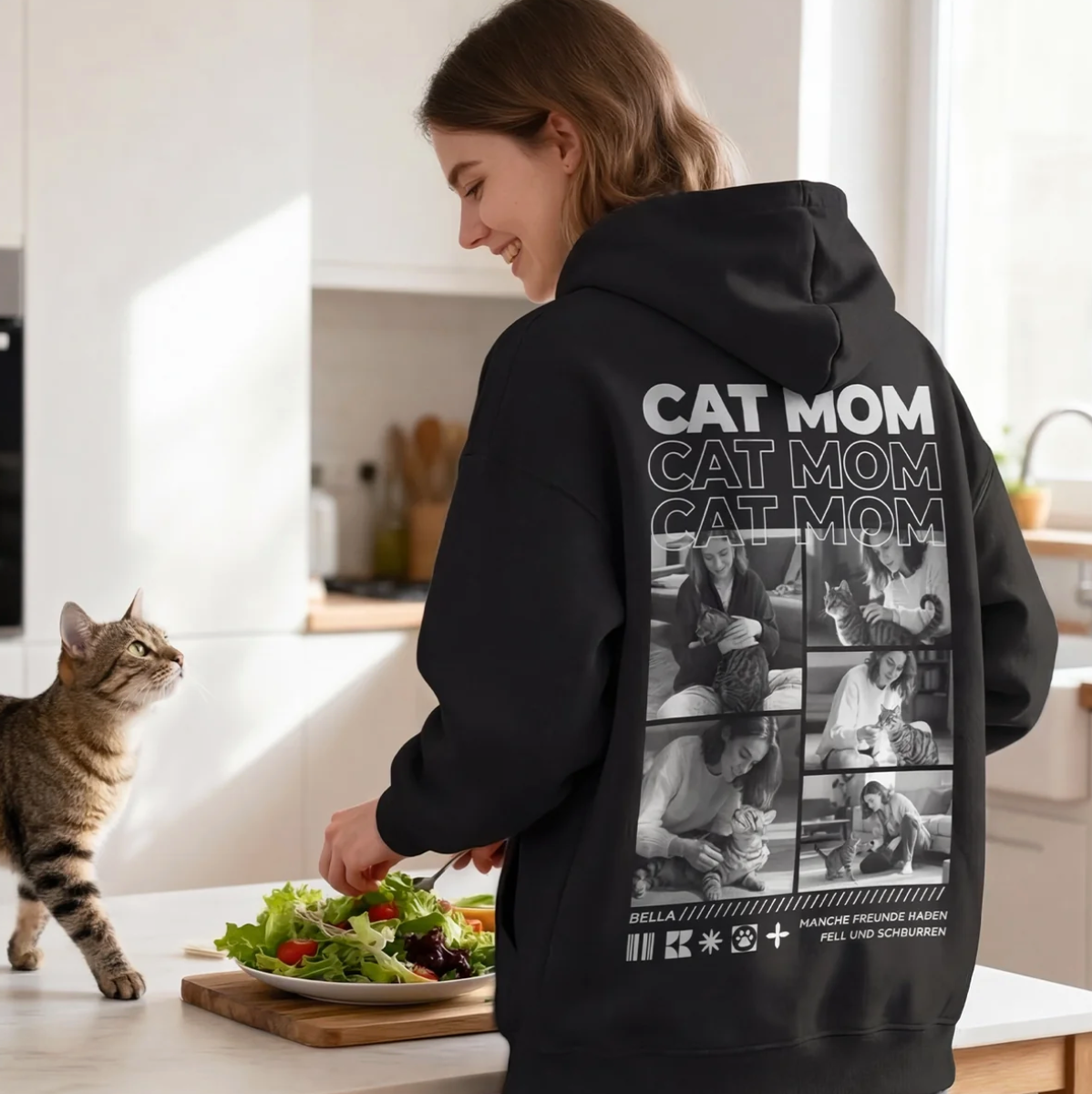 Schwarzer Hoodie mit "Cat Mom"-Print auf dem Rücken, Person steht in der Küche neben einer Katze.