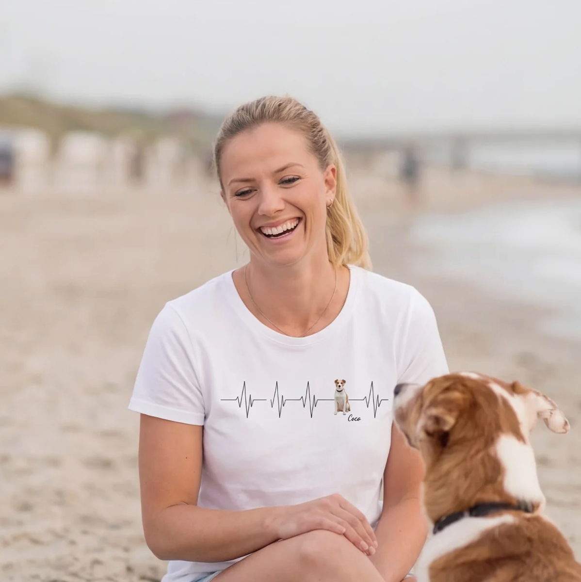 Weißes T-Shirt mit Herzschlag-Muster und Hundemotiv, sitzende Frau am Strand.