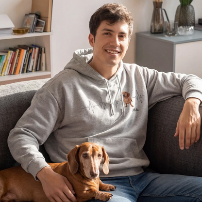 Grauer Hoodie mit Herzschlagmotiv und Hundebild, Träger sitzt auf einer Couch mit Dackel im Arm.