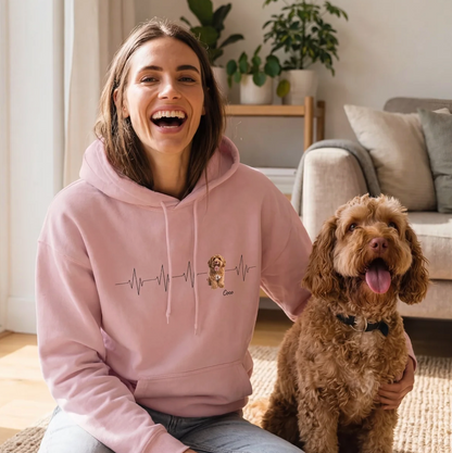 Person tragt einen rosa Hoodie mit Herzschlag-Motiv, sitzend auf einem Teppich neben einem Hund.