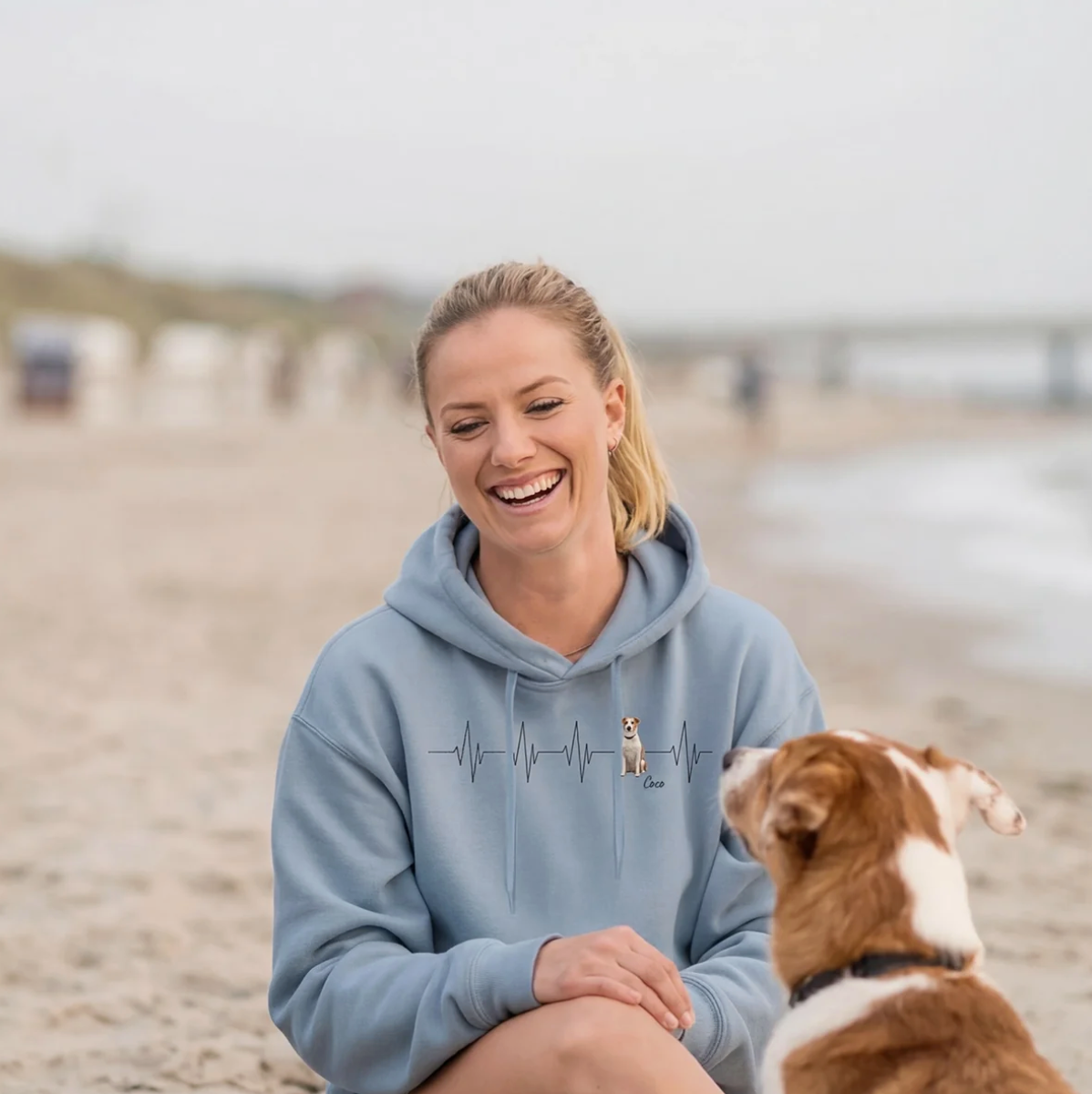 Personalisierter blauer Hoodie mit Herzschlag-Motiv und Hundemotiv, Frau sitzt am Strand und lächelt.
