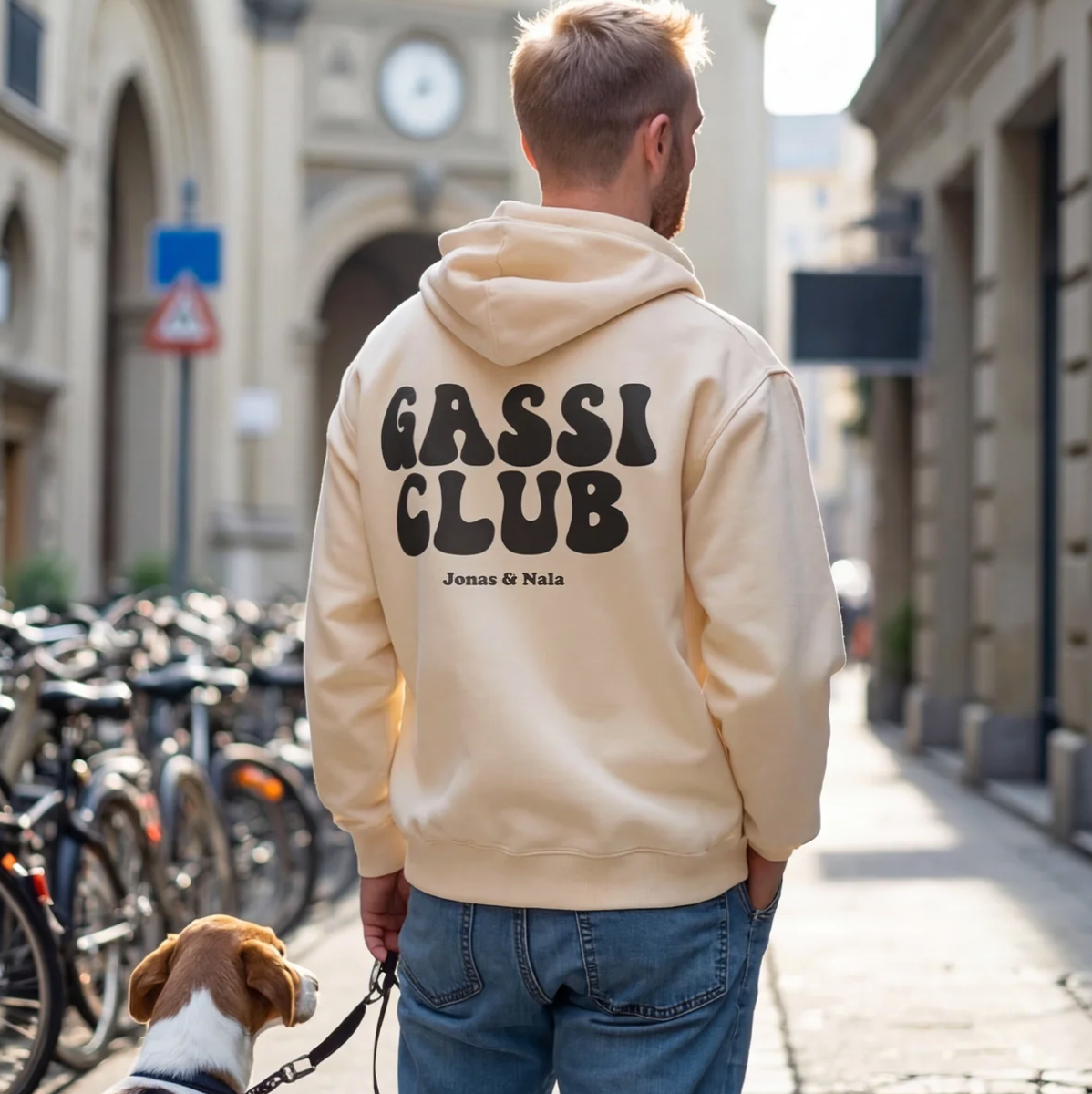 Person trägt einen beigen Hoodie mit der Aufschrift "Gassi Club" hinten, steht mit Hund in einer Stadtstraße.