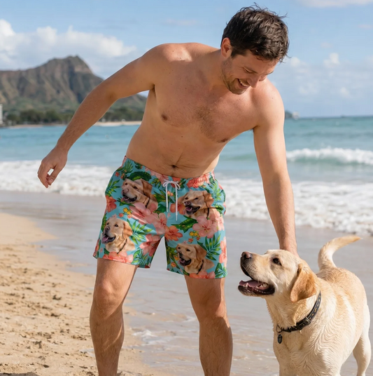 Mann am Strand trägt bunte Badehose mit Hundemotiv und Blumen. Labradorhund spielt neben ihm im Sand.