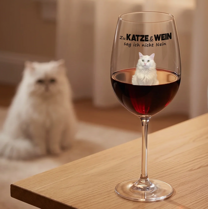 Personalisiertes Weinglas mit schwarzer Schrift und rotem Wein, daneben eine weiße Katze auf einem Holz Tisch.