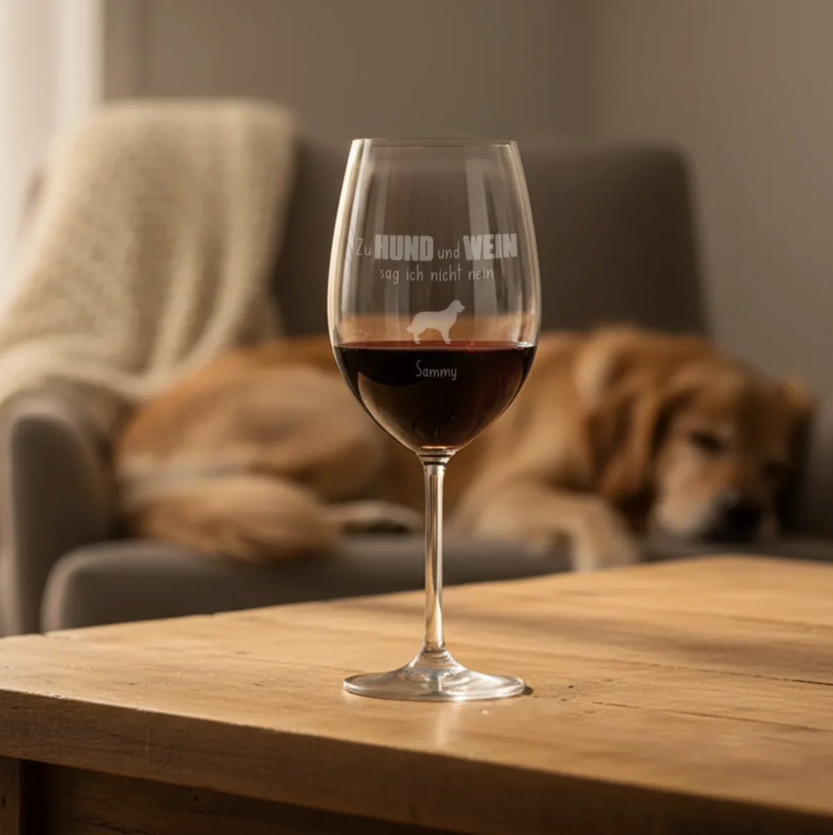 Personalisiertes Weinglas mit Gravur, gefüllt mit Rotwein, im Vordergrund auf einem Holztisch. Hintergrund mit Hund.