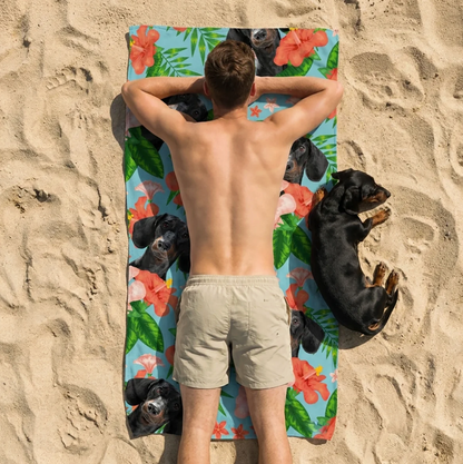 Person liegt auf einem personalisierten Strandtuch mit Hundemotiven, umgeben von Sand.