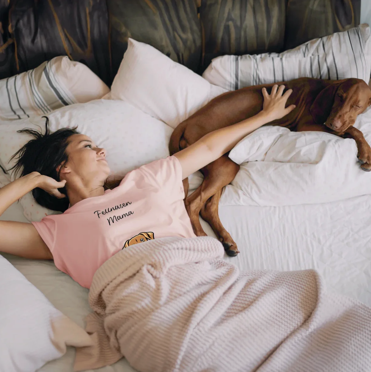 Frau in pinkem T-Shirt liegt auf dem Bett neben einem Hund, umgeben von Kissen und einer Decke.