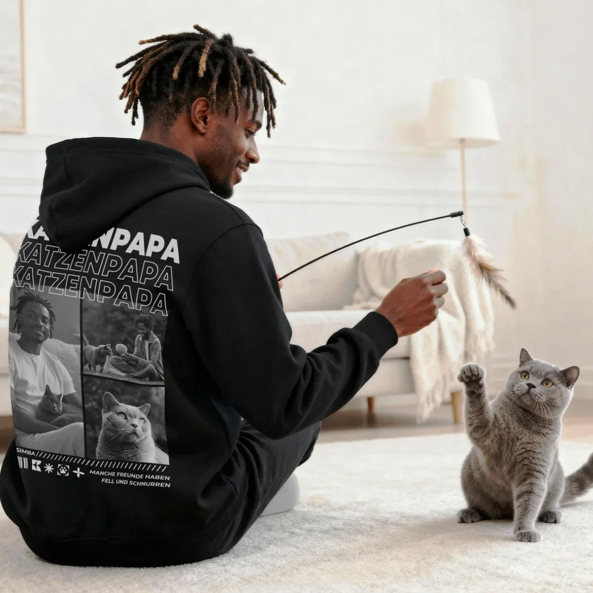 Person spielt mit grauem Kater, trägt schwarzen Hoodie mit Katzenmotiven. Helle, moderne Wohnumgebung.