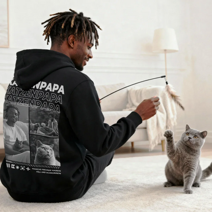 Person spielt mit grauem Kater, trägt schwarzen Hoodie mit Katzenmotiven. Helle, moderne Wohnumgebung.