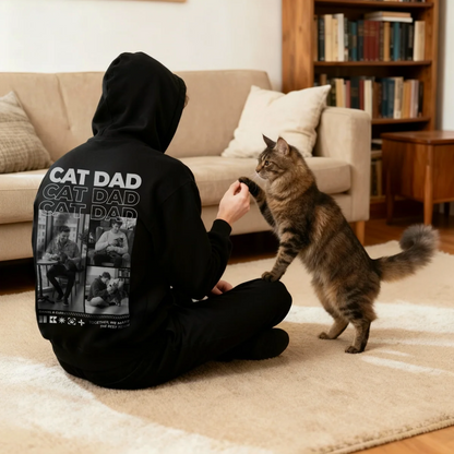 Schwarzer Hoodie mit "Cat Dad"-Aufdruck auf dem Rücken, Person sitzt auf dem Boden und spielt mit einer Katze.